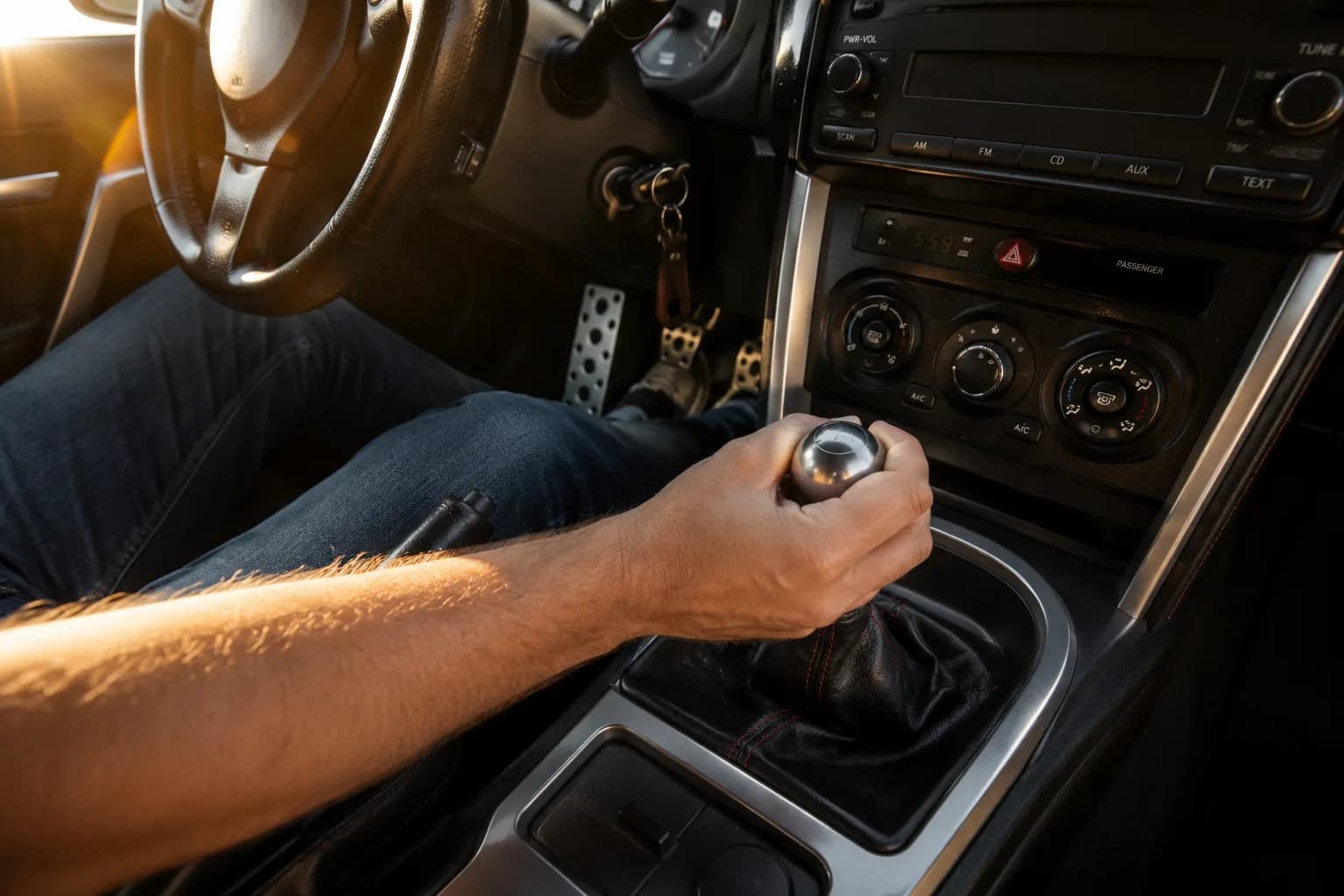 Close up view inside a classic sports car showing a driver's hand on a leather-wrapped manual gear shifter with three pedals visible in the footwell.
