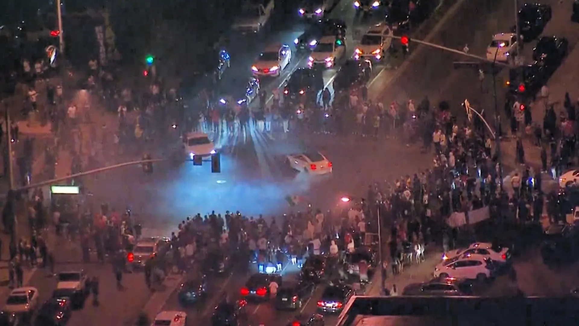 Aerial nighttime view of an illegal street takeover with cars doing donuts in a blocked intersection surrounded by a large crowd.